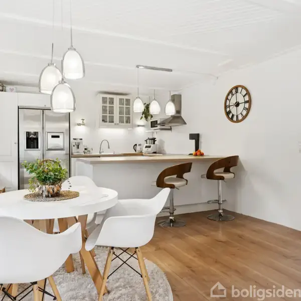 Et lyst, moderne køkken med hvidt spisebord og stole i forgrunden. Køkkenø med barstole til højre. Omkring er der trægulve, hvide skabe, loftslamper og en vægur.