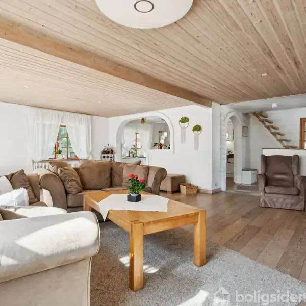 Stue med lysebrune sofaer og sofabord i træ. Trappen fører til en øverste etage. Rummet har trægulv, hvide vægge, store vinduer, og planter, hvilket skaber en lys atmosfære.