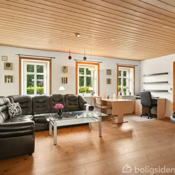 Sort lædersofa står foran et glasbord med en vase med blomster. I baggrunden er et skrivebord med stol foran tre store vinduer, der lukker naturligt lys ind.