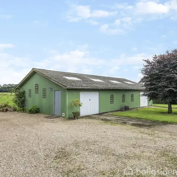 Grøn bygning står stille ved grusvej i landligt område. Store vinduer og en hvid dør er synlige. Et stort træ og marker omgiver bygningen under en blå himmel.