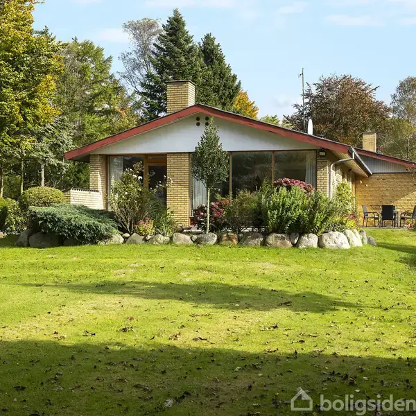 Huset står midt i en velholdt have med græsplæne og buske, omgivet af træer. Det har et skråt tag, en skorsten og store vinduespartier.