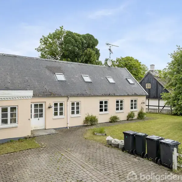 En lang, lyserød bygning med flere vinduer og en dør. Taget har flere ovenlysvinduer. Omgivet af en brostensbelagt gårdsplads og grønne buske.