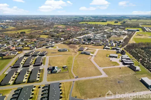 En gruppe huse står stille i en nybygget boligområde omgivet af åbne marker og veje i forgrunden, med en by i horisonten under en skyfri himmel.