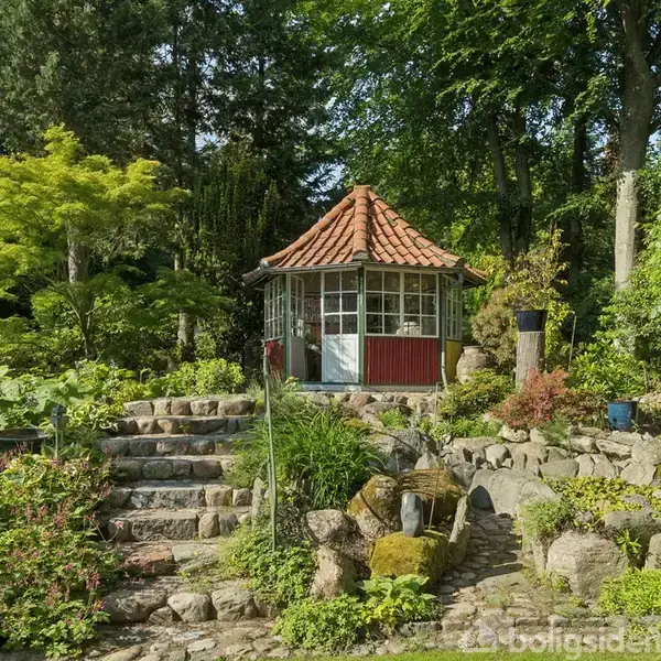 Pavillon står stille i en frodig have omgivet af træer og buske, beliggende ved en stenbelagt trappe.