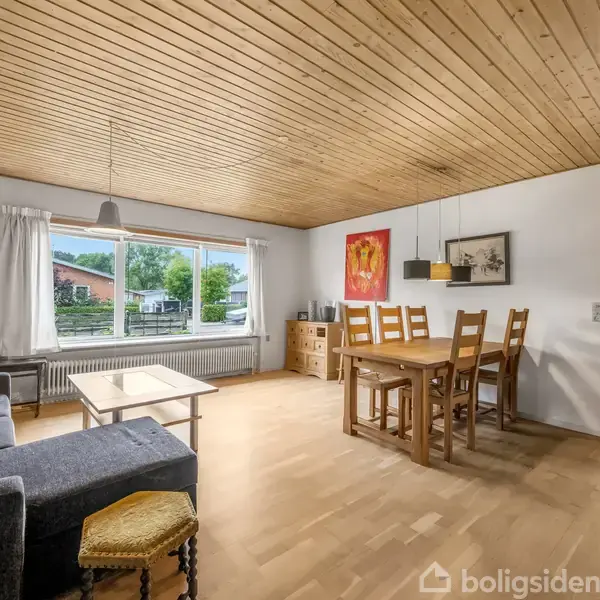 Stue med træloft og parketgulv, indeholder en grå sofa, et sofabord, spisebord med seks stole. Store vinduer viser en have udenfor. Væggene prydes med forskellig kunst.