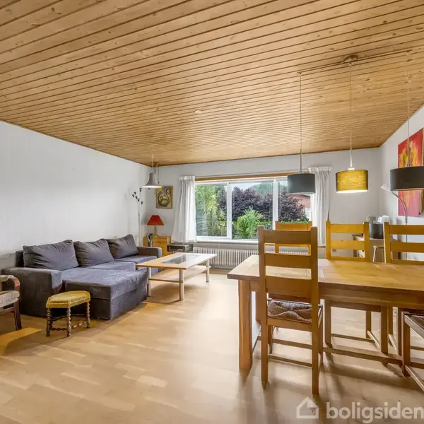 Sofa med puder står ved vindue, ved siden af lænestol og småborde. Spisebord med stole ved siden af. Loftslamper hænger ned. Væggene har billeder. Trægulv og træloft.