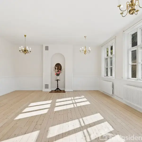 Et lyst, tomt rum med trægulv og to lysekroner hængende fra loftet. En vase med blomster er placeret i en indretningsniche. Tre store vinduer til højre.