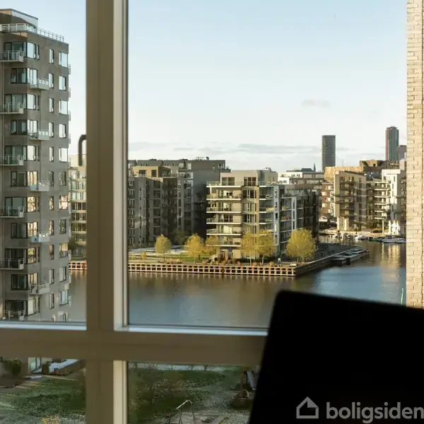 En udsigt fra et vindue viser høje, moderne lejlighedsbygninger omkring en kanal med træer og vand. Skyer ses på himlen, og en laptop anes i forgrunden.