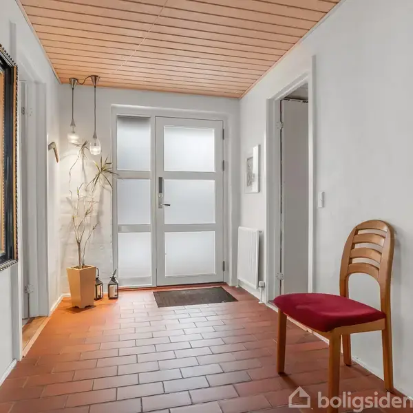 En træstol med rødt sæde står i en entre. En plante og lanterner står ved døren, som har frostet glas. Et stort spejl hænger på væggen ved siden af.