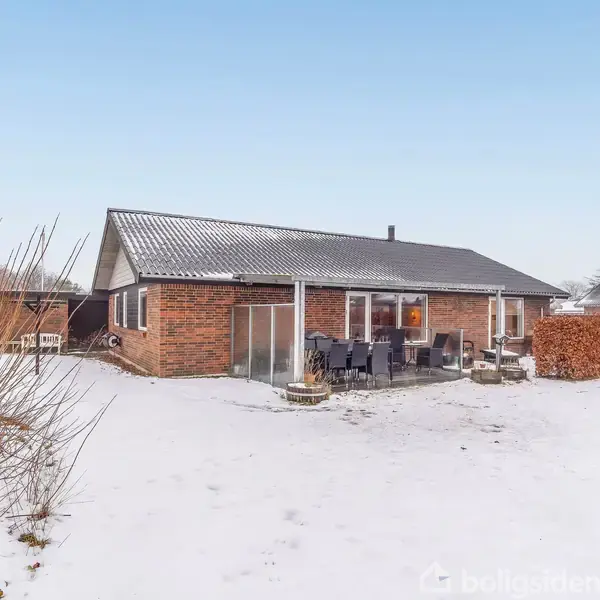 Et murstenshus står i en snedækket have. Der er havemøbler på en terrasse foran huset. Omkring er der buske og træer under en klar himmel.