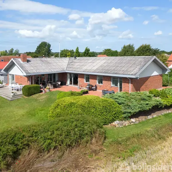 En étplans rødstensvilla med terrasse og have omkranset af buskads og græsplæne. Det er solrigt ude med flere naboer, der har tilsvarende terracottafarvede tagsten. Himmel med spredte hvide skyer.