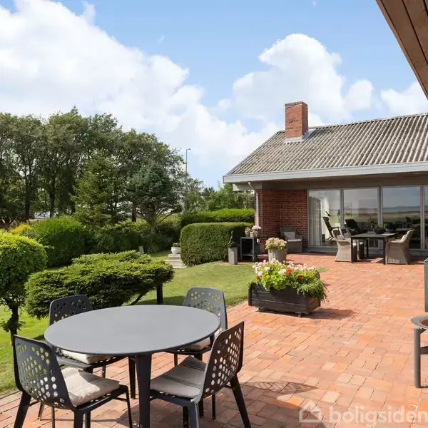 Rundt havebord står på en terrasse lavet af røde mursten; omkranset af stole og planter. I baggrunden er et rødt murstenshus med store vinduer og grønt haveområde.