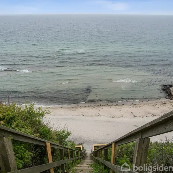 Trappe fører ned til en sandstrand ved kysten. Havet ses i baggrunden med små bølger, omgivet af grøn vegetation på klitterne.