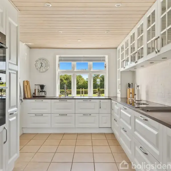 Et lyst køkken med hvide skabe og mørke bordplader. Vindue med udsigt til grønne omgivelser. Der er en indbygget ovn og forskellige køkkenredskaber på bordet. Vægur hænger til venstre.