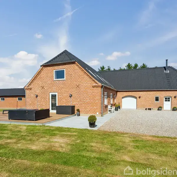 Murstenshus med sort tag står på en åben grusvej i landligt område med træer i baggrunden og himmel over. Terrasse med havemøbler og græsplæne foran.
