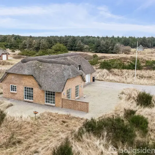 Et stråtækt hus står stille på en flisebelagt grund omgivet af klitlandskab og græsser. En bil er parkeret til højre, og der ses træer i det fjerne.