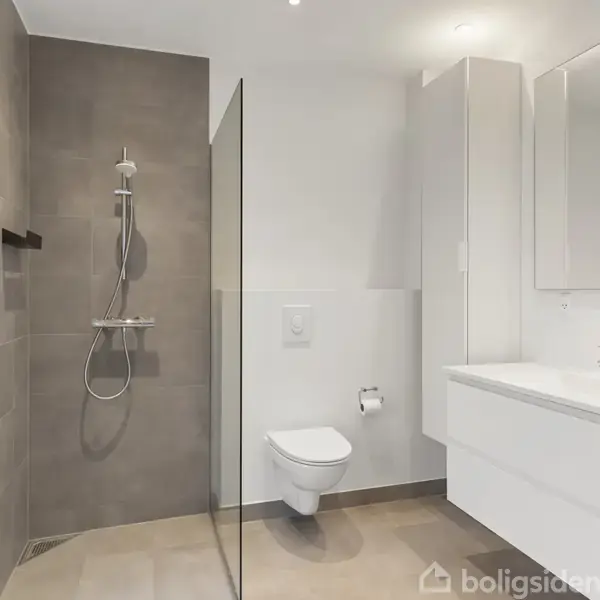 Brusebadet er adskilt af en glasvæg, der står ved siden af et væghængt toilet. Ved spejlet og vasken ses en hvid vaskemaskine i en lys og moderne badeværelsesindretning.