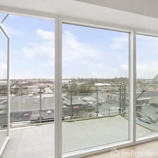 En stor glasdør står åben mod en balkon. Udenfor ses en byudsigt med bygninger i baggrunden under en klar blå himmel.