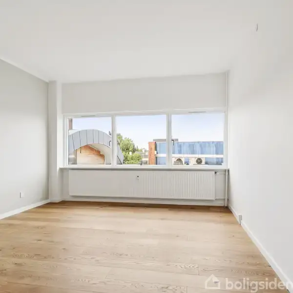 Et lyst, tomt værelse med lyse trægulve og hvidmalede vægge. Store vinduer giver udsigt til bygninger udenfor. Radiator placeret under vinduerne.