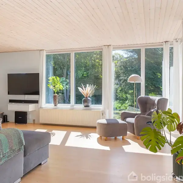 Stue med grå sofa, pejs og stor fladskærm. Store vinduer lader naturligt lys strømme ind, mens grønne planter tilføjer liv. Lyse træmøbler og gulv skaber en åben, indbydende atmosfære.