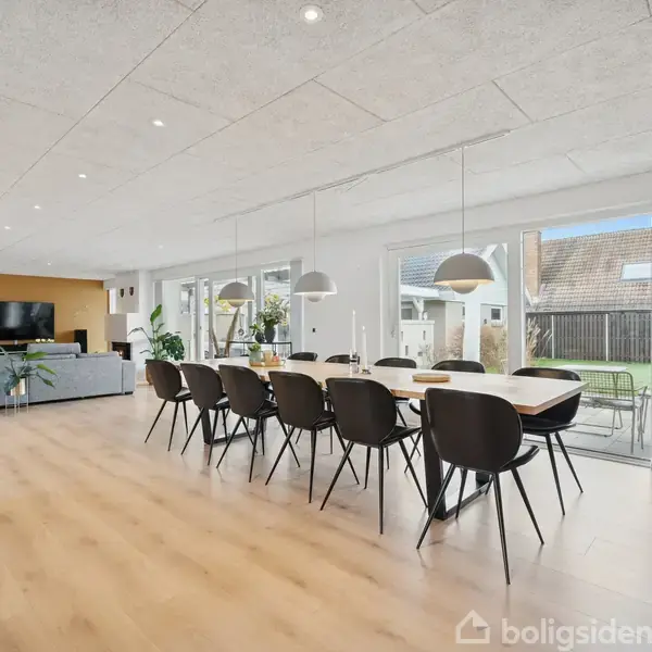 Spisebord med sorte stole står i en lys stue, hvor store vinduer vender ud mod en terrasse. Baggrunden viser en opholdsstue med sofa, tv og planter.