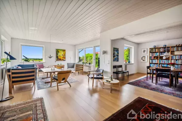 Stue med moderne møbler; lyse trægulve og store vinduer, der giver udsigt til grønne omgivelser. Bogreol fyldt med bøger i tilstødende rum. Malerier hænger på væggene.