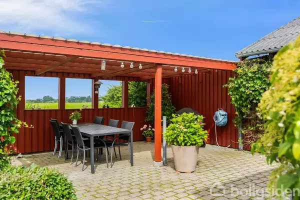 Sort spisebord med stole står under en åben, rød træpergola. Omgivet af grønne planter og fliser, ses marker og himmel i baggrunden. En haveslange hænger på væggen.