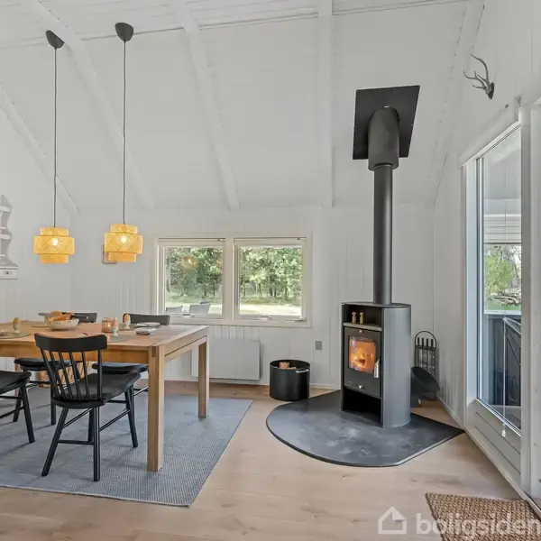 Brændeovn står ved siden af et vindue og udsender varme, mens en træspisebord med sorte stole er placeret under to gule lamper. Området er lyst og hyggeligt.
