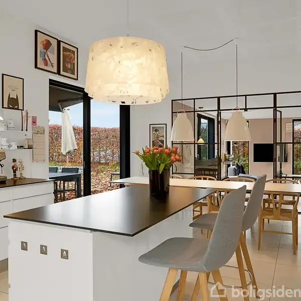 Køkkenø med to stole står i et moderne køkken. Der hænger en stor lampe over øen. Baggrunden indeholder et spisebord og vægdekorationer. Vaser med blomster pynter bordet.