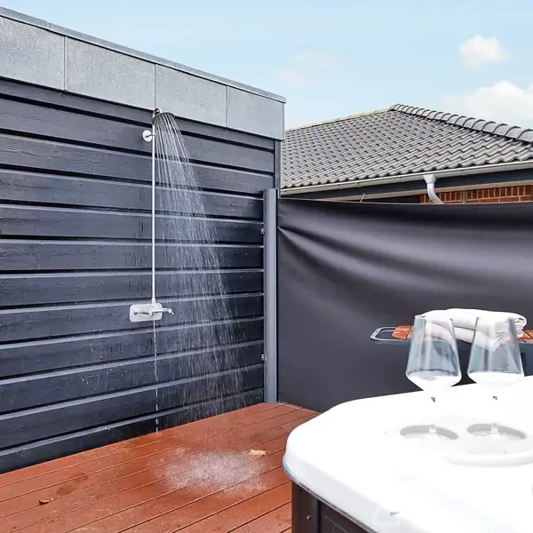 Udendørs bruser hænger på en moderne, sort trævæg og sprøjter vand. Ved siden af står et spa-kar med to vinglas og en uåbnet champagneflaske. Hustag ses i baggrunden.