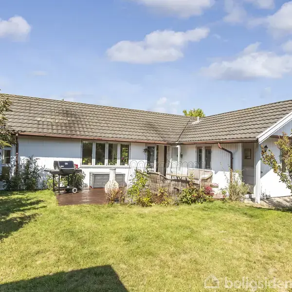 Hus med grå tag, placeret i en grøn have. En grill og terrasse er foran huset, omgivet af buske og træer. Hvide skyer fylder den blå himmel.