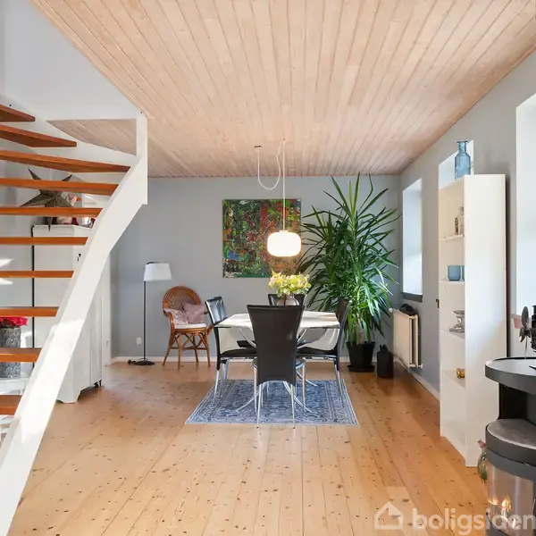 Trætrappe fører opad, mens spisebord med sorte stole står nedenunder. Lampen hænger over bordet. Stuen har lyse trælofter, store grønne planter og en rund brændeovn i hjørnet.