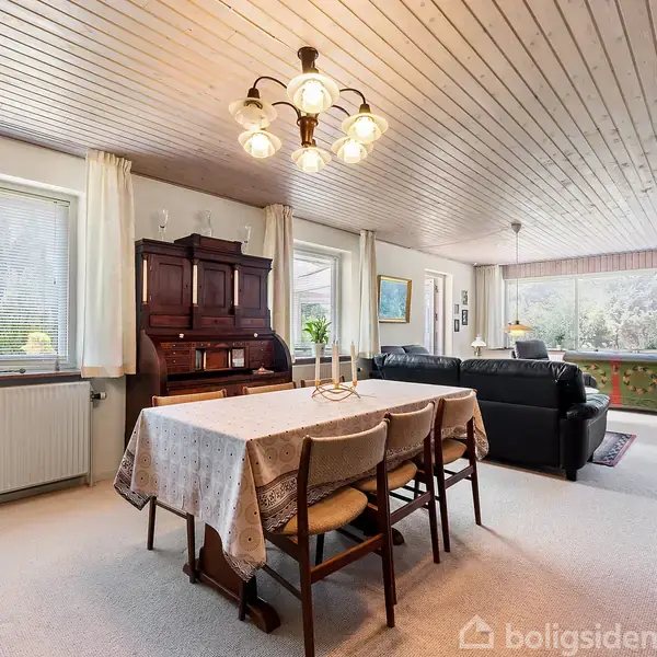 Et spisebord med dug og stole står midt i en stue. Rummet har en sofa, et klaver, store vinduer og et reolsystem, som skaber en lys og hyggelig atmosfære.
