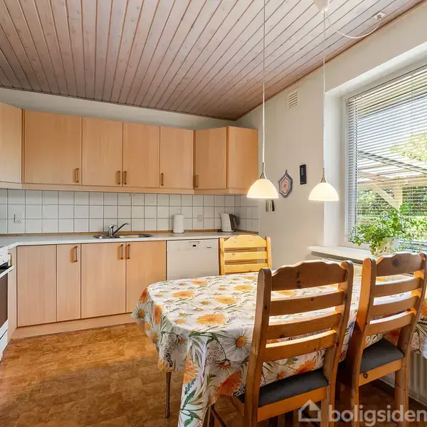 Køkken med lyse træskabe arrangeret i en vinkel, en ovn og en mikrobølgeovn på bordpladen. I midten står et bord dækket med en blomsterdug, omgivet af træstole.