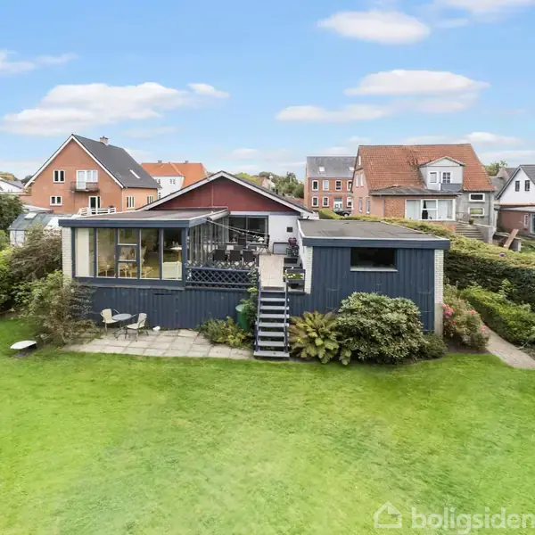 Blåt hus står på en grøn græsplæne med trin og terrasse, omgivet af buske. I baggrunden ses andre huse og en blå himmel med få skyer.