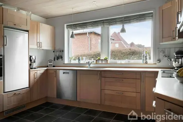 Et rummeligt køkken med lyse træskabe og grå flisegulv har en stor vinduesvæg. Udsigt til en nabobygning gennem vinduet, med planter på vindueskarmen. To lamper hænger over vasken.