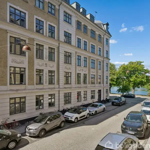 Bygning med flere etager står langs en brostensvej, hvor biler er parkeret. Træer og en sø er synlige i baggrunden under en klar blå himmel.