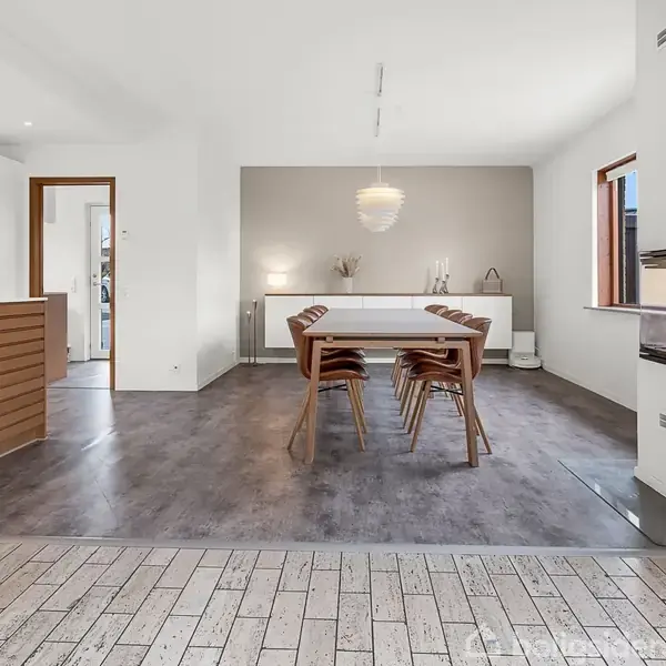 Et spisebord med seks stole står i et lyst spiseområde med trælaminatgulv. Ved siden af er der et moderne køkken med hvide elementer og en delvist åben murstensvæg.