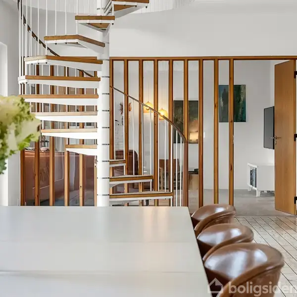 En spiraltrappe fører opad i en moderne stue med trægulve. Et hvidt spisebord står i forgrunden med en buket hvide blomster, flankeret af brunlæderstole.