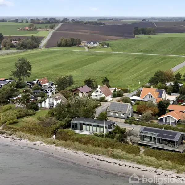 Kysthuse ligger langs en strand. Huse med forskellige tagfarver er omgivet af træer. Bag husene strækker åbne marker sig mod horisonten.