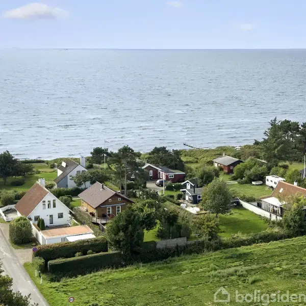 Huse ved kysten, nogle med røde tage, omgivet af grønne haver og træer. Vejen løber parallelt med havet, der strækker sig til horisonten under en blå himmel.