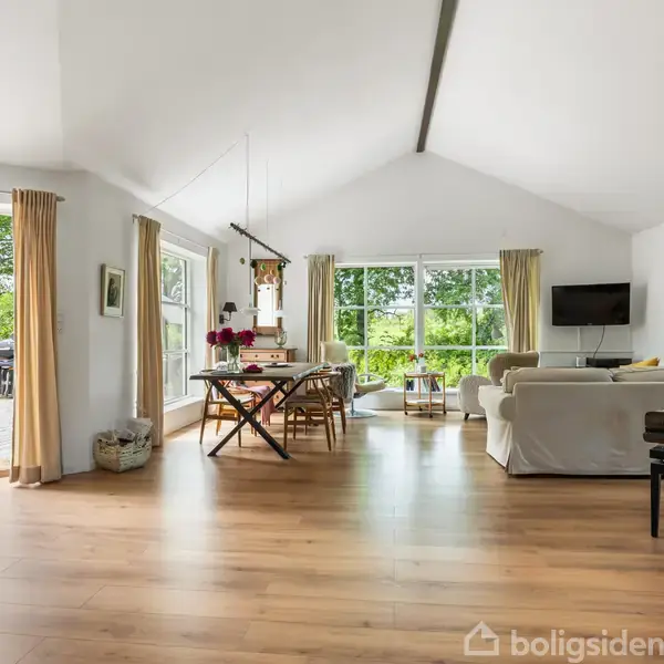 Stue med trægulv, hvidmalede vægge og loft. Møblerne inkluderer sofaer, spisebord og flygel. Store vinduer slipper lys ind og viser en terrasse udenfor med borde og grønt.
