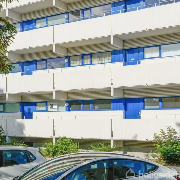 En flerfamiliebygning med blå døre og vinduer. Biler holder parkeret foran bygningen, og der er grønt buskads i forgrunden.