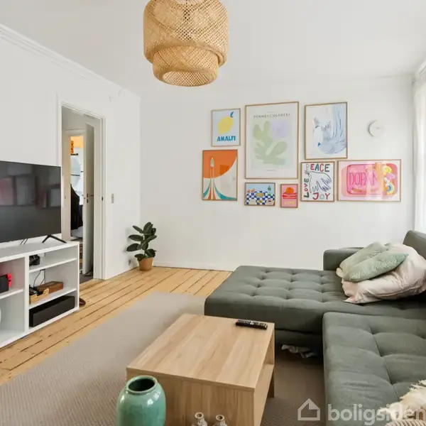 Grøn sofa står i en lys stue med trægulv, flankeret af et sofabord og tv. Væggene prydes af farverige plakater. En indretningsplante står i hjørnet.