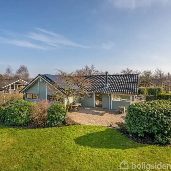 En blåmalet enfamilieshus med sort tag står omgivet af en velplejet have. Sollys skinner ned på græsplænen, mens buske og træer dekorerer området omkring huset.