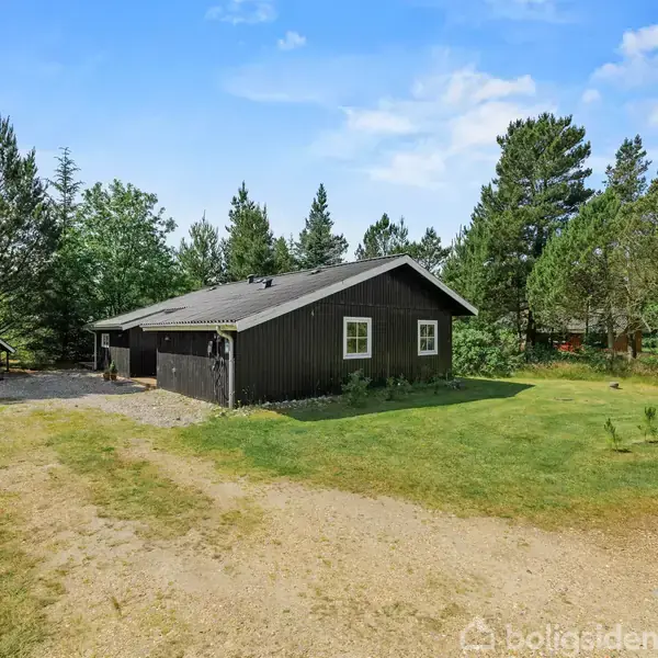 Et sort træhus står stille på en grusbelagt grund, omgivet af græsplæne og tætte skove under en klar blå himmel.