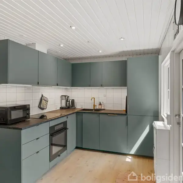 Køkken med grønne skabe, mikroovn og ovn på venstre side. Lys træbordplade med hvid flisevæg. Store vinduer giver rummet lys. Indbydende opstilling i et moderne hjem.