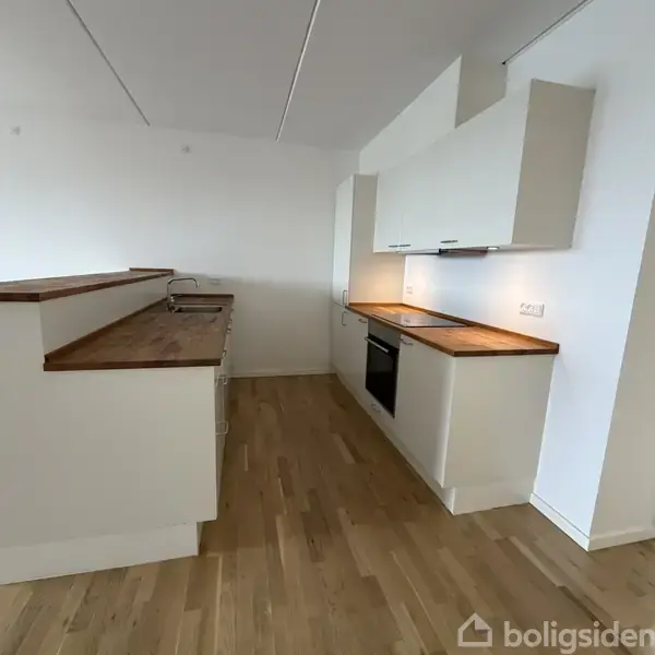 Et moderne køkken med hvide skabe og træborde. Ovnen og kogepladen er indbygget på højre side, mens vasken er til venstre. Rummet har trælaminatgulv.