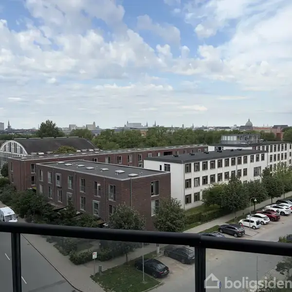 Flere sammenkoblede bygninger står stille omgivet af træer, med grå skyer let spredt over himlen som en baggrund, indikerende et byområde.