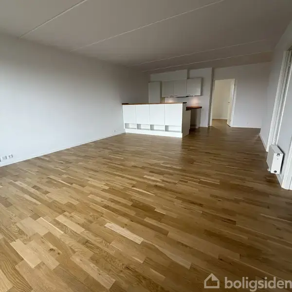 Stue med lyst trægulv, åben mod et lille køkken med hvide elementer og en bar. I baggrunden ses en døråbning til et tilstødende rum.
